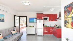 A modern living room with a grey sofa and a flat-screen TV. A kitchen with red cabinets and a white refrigerator is visible.