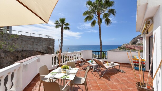 A terrace with a table set for dining, overlooking a coastal area with palm trees and a view of the sea.