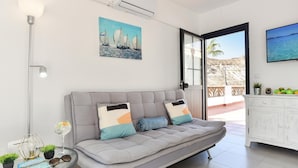 A modern living room with a grey sofa, a glass coffee table, a painting of sailboats, and a view of the sea through an open door.