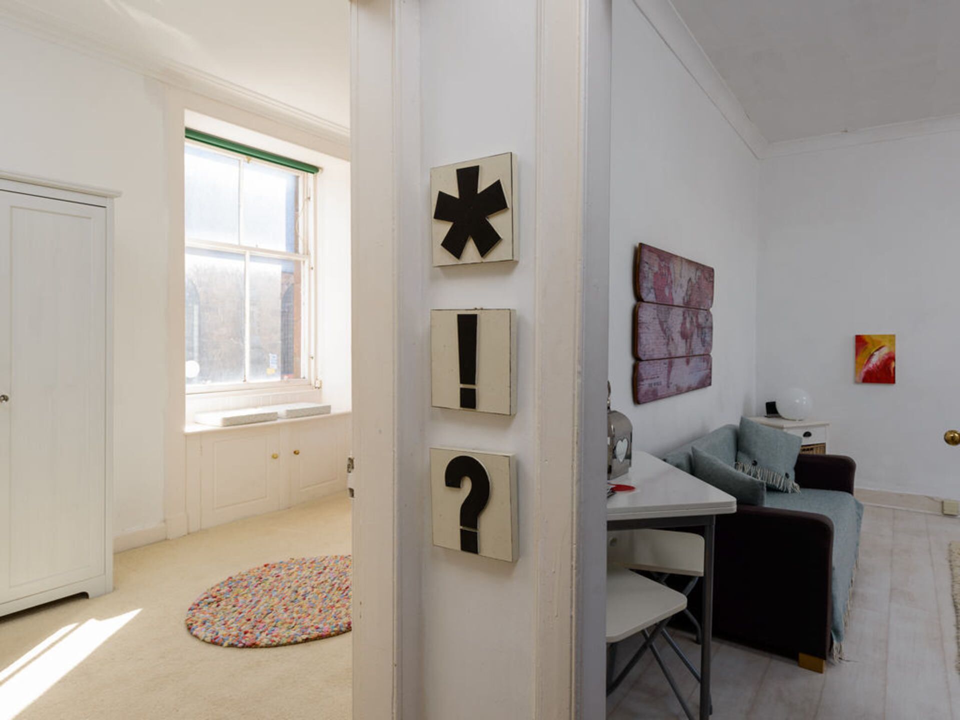 A well-lit room with a white door, a window with green trim, a white table, a black chair, a small rug, and a wall with three black and white signs.