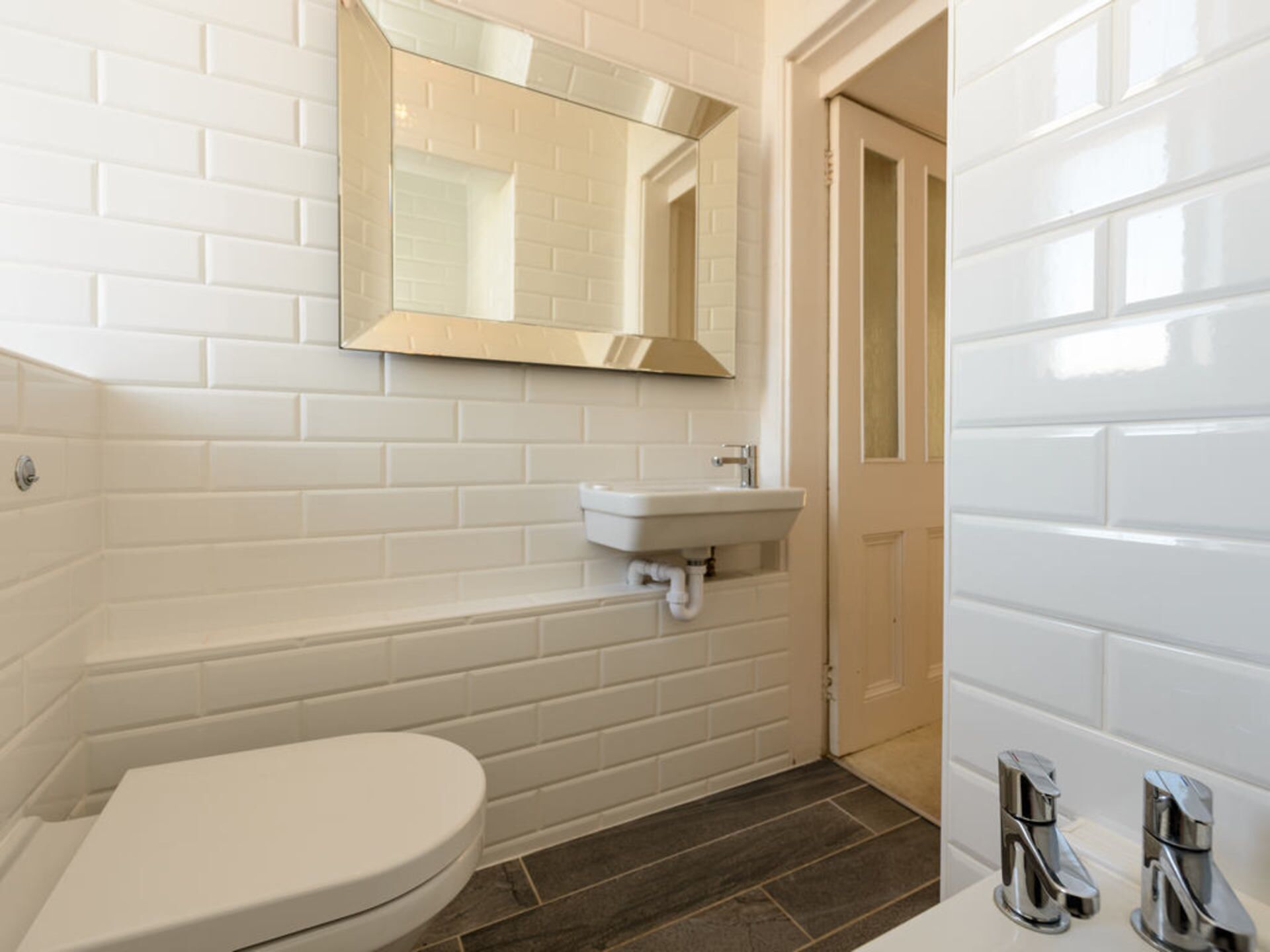 Un bagno con piastrelle bianche, un water bianco, un lavandino bianco con uno specchio sopra e una zona doccia.