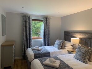 A bedroom with two beds, patterned curtains, a bedside lamp, and a view of greenery outside.