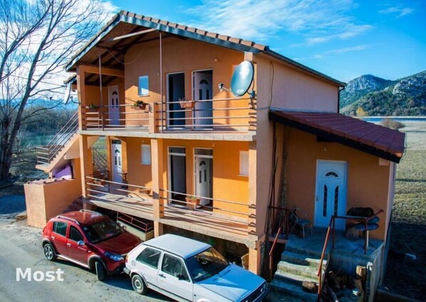 Two-story building with a balcony and flat roof, set amidst a natural landscape with trees and mountains.