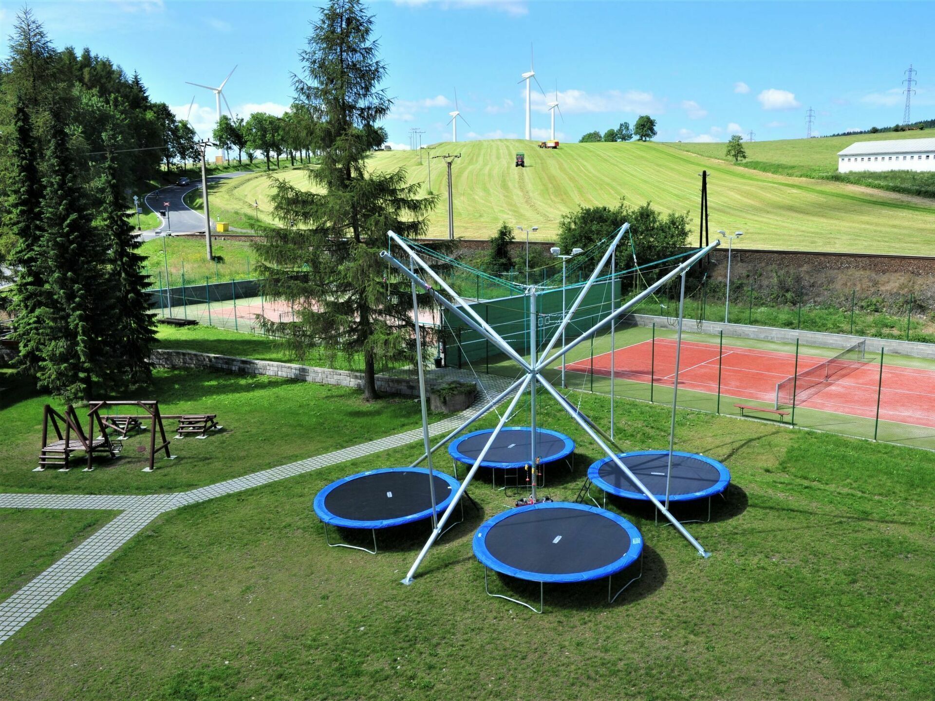 A playground with multiple trampolines, a tennis court, and wind turbines in the background.