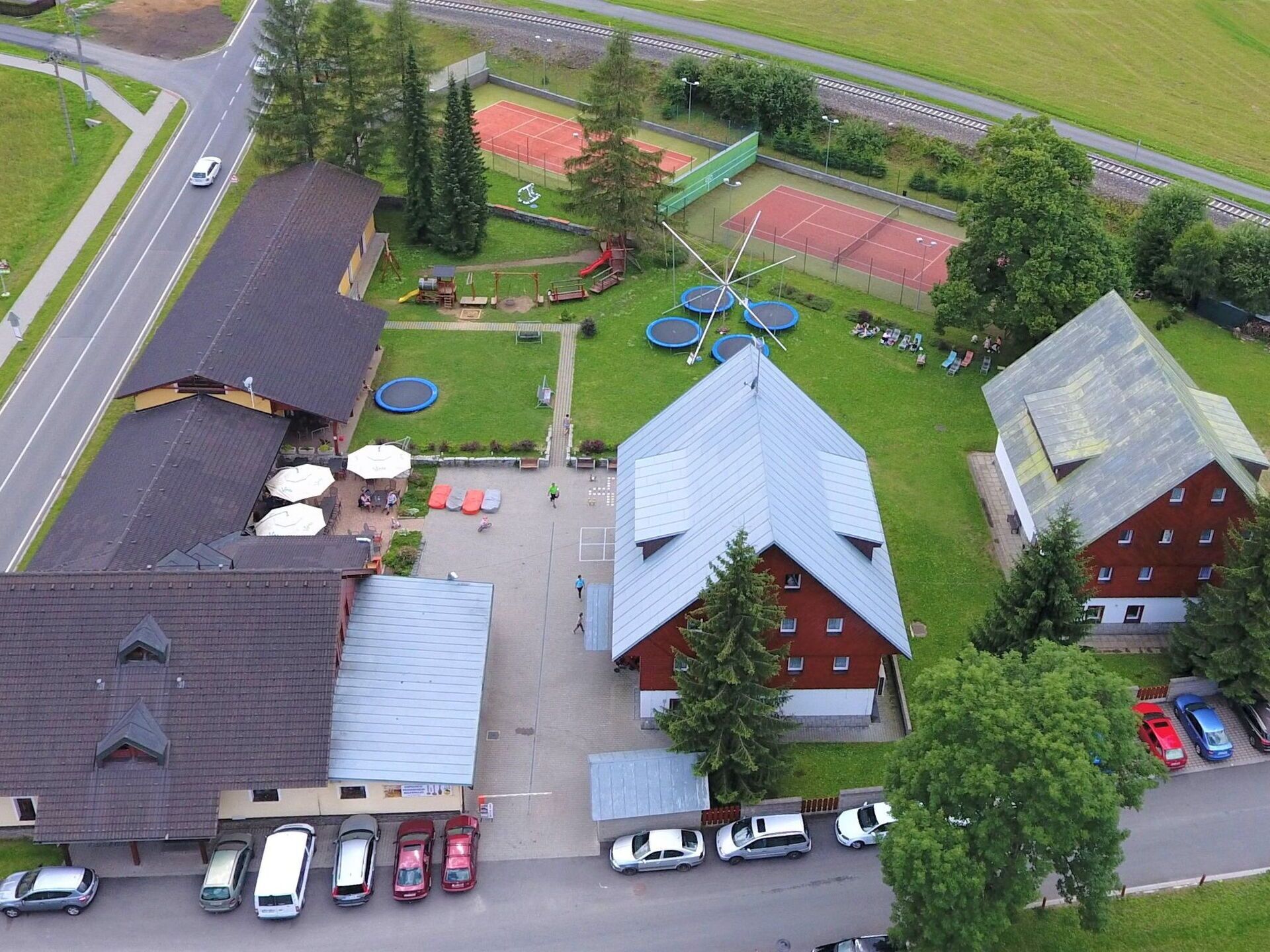 An aerial view of a building with a sloped roof, a parking lot with several cars, and a tennis court with a net.