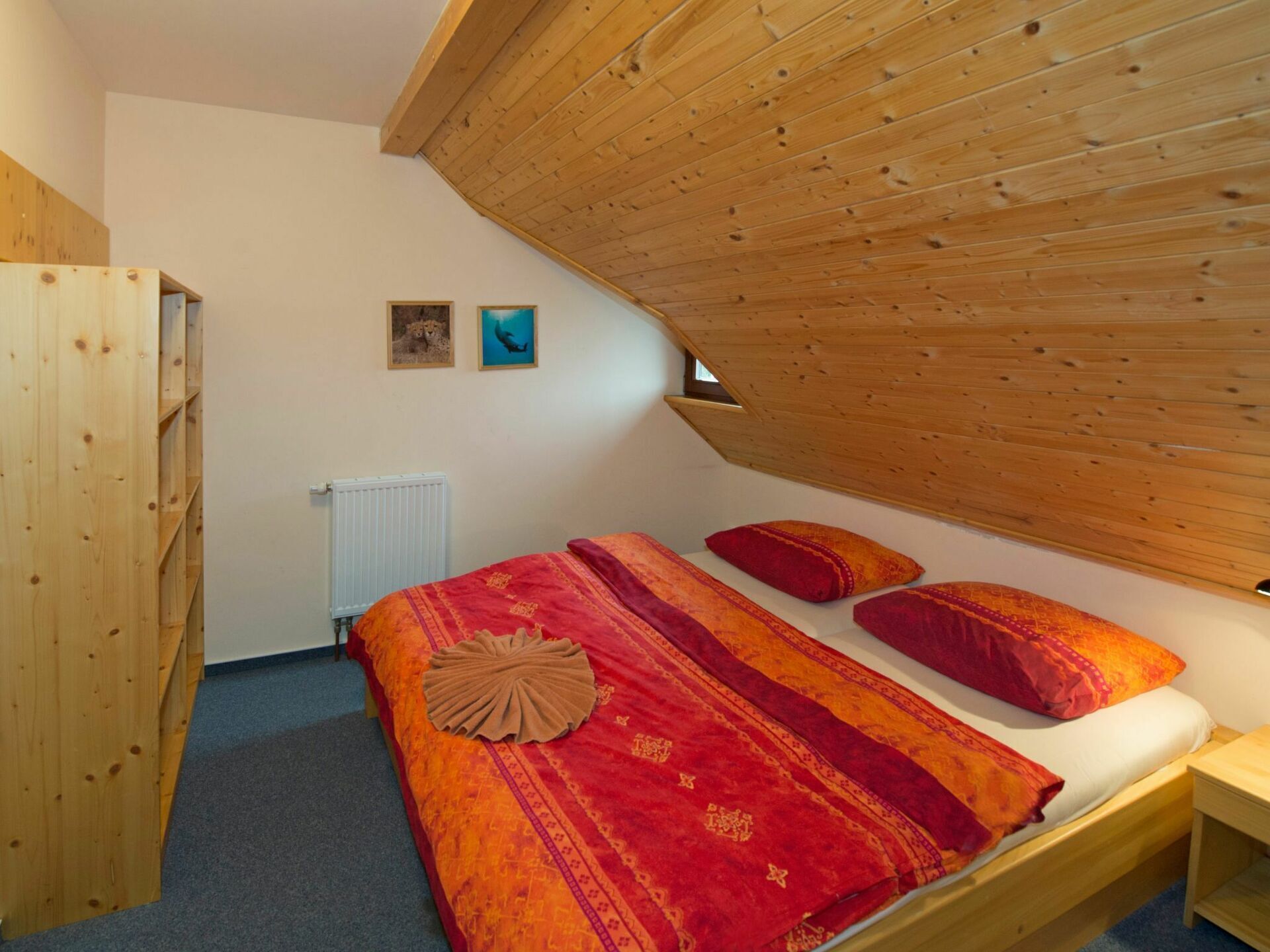 A bedroom with a sloped ceiling, a bed with red bedding, two pillows, a wooden wardrobe, and a radiator.