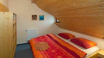 A bedroom with a sloped ceiling, a bed with red bedding, two pillows, a wooden wardrobe, and a radiator.