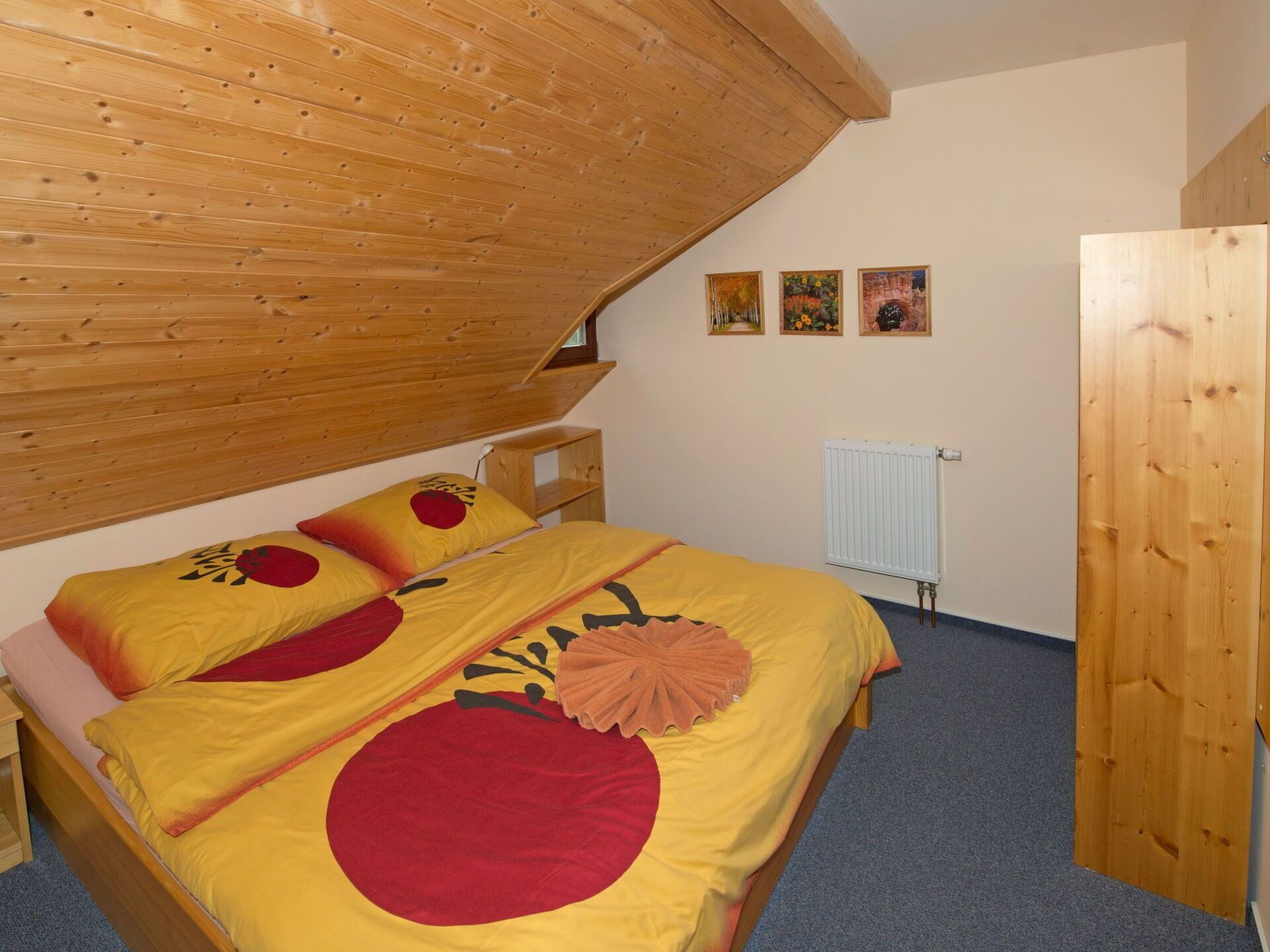 Une chambre avec un plafond en pente, des murs en bois, un lit avec une literie jaune et rouge, une petite table de chevet et un radiateur.