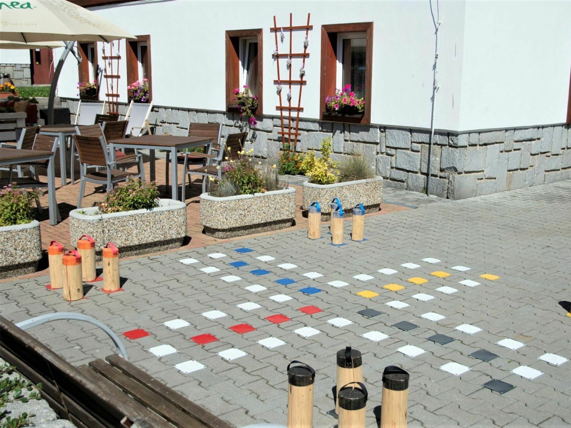 An outdoor seating area with tables and chairs, planters with flowers, and a paved ground with colorful tiles.