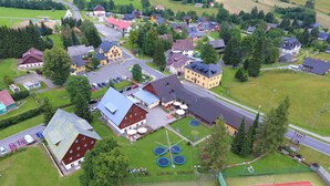 An aerial view of a village with a central building surrounded by houses, a tennis court, and a parking area.