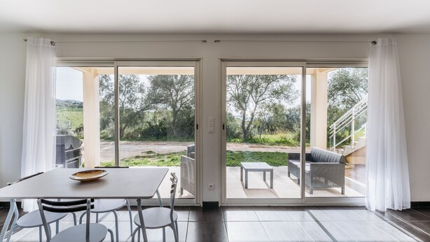 Ein modernes Zimmer mit einer Schiebetür zu einer Terrasse. Das Zimmer verfügt über einen Esstisch mit Stühlen, ein Sofa und einen kleinen Tisch mit einer Bank.