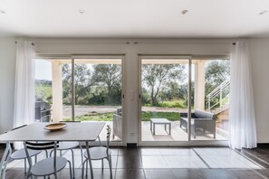 A modern room with a sliding glass door leading to an outdoor patio. The room has a dining table with chairs, a sofa, and a small table with a bench. - Matteo Leccia - appartement avec piscine (Peri)