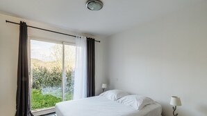 Ein Schlafzimmer mit Bett, Nachttisch, Lampe und Blick auf Grün hinter einem Fenster.