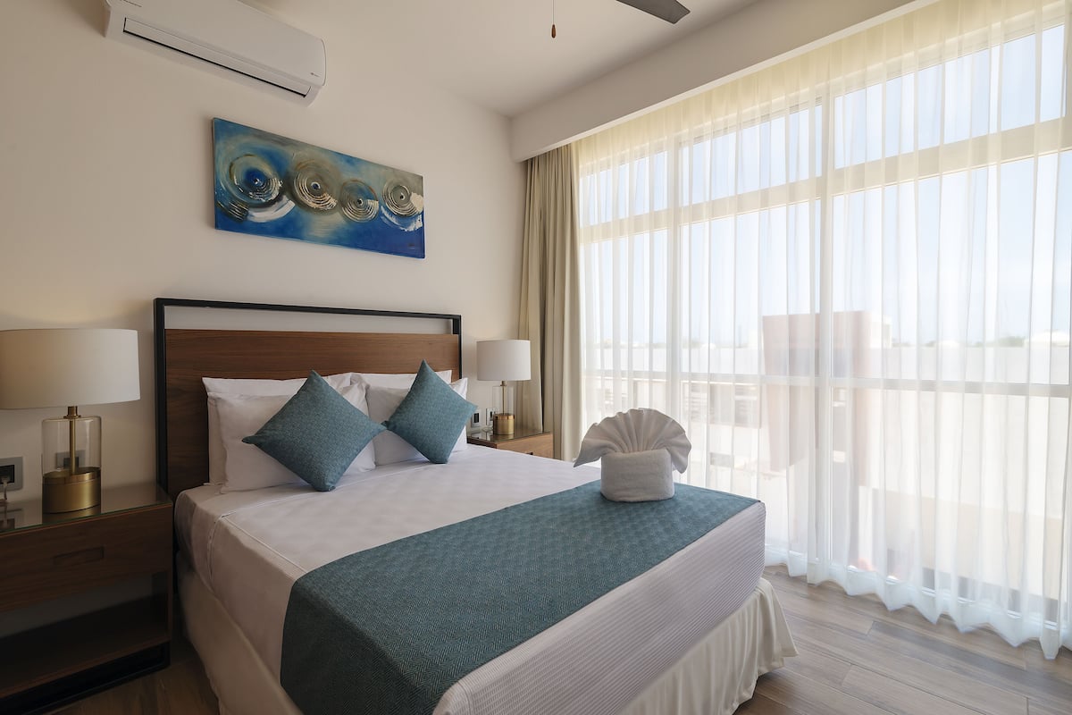Een hotelkamer met een groot bed, twee nachtkastjes met lampen, een schilderij aan de muur en dunne gordijnen.