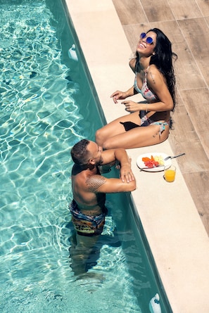 A man and a woman sitting by a swimming pool, with a plate of food and a drink on the poolside.