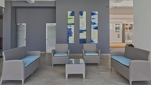 Uma área de recepção com três sofás de vime e uma mesa de centro de vidro, parede cinza com arte abstrata, e vista para o lobby.