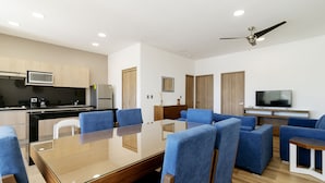 Uma cozinha moderna com micro-ondas, fogão e frigorífico. Possui uma área de jantar com mesa de vidro e cadeiras azuis. É visível um ventilador de teto, e uma televisão está fixada na parede.