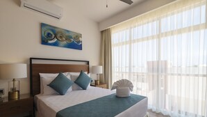 Cama bem arrumada com colcha verde-água, duas almofadas e uma toalha branca ao lado. Há um quadro na parede acima da cama, uma mesa de cabeceira com abajur e uma janela com cortinas leves.