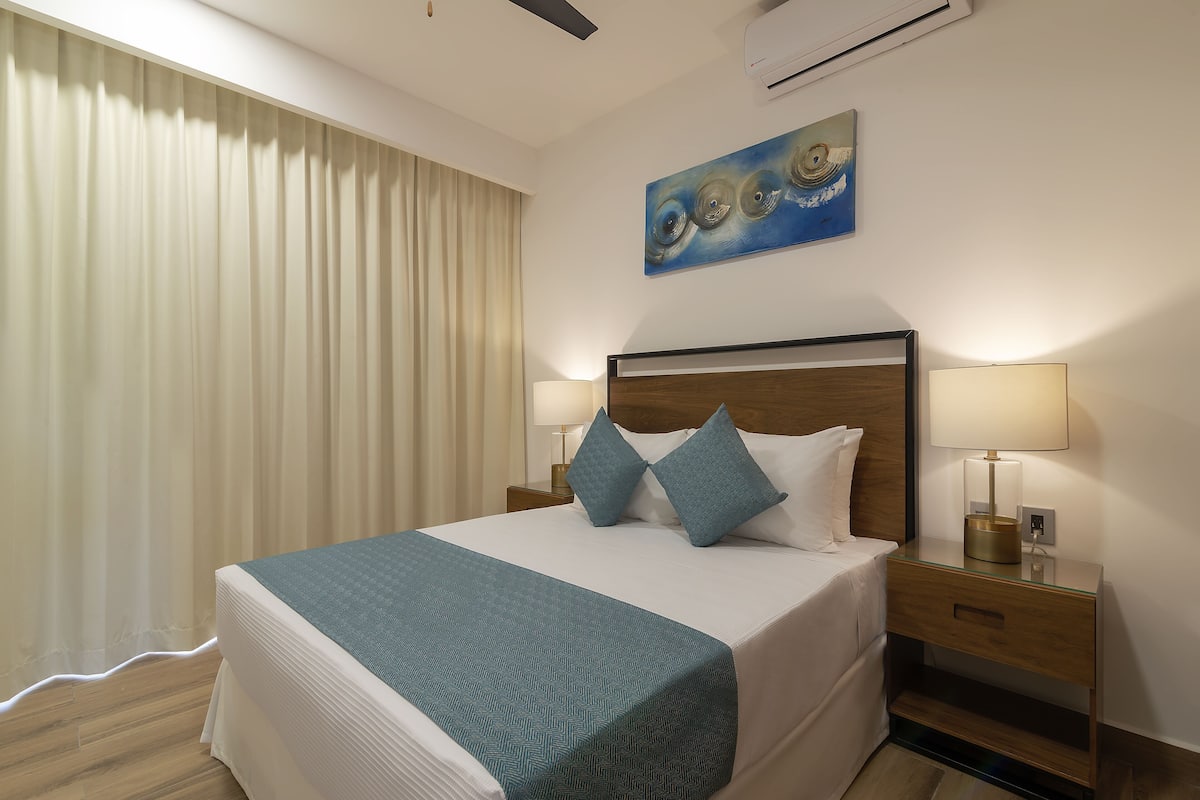 Een hotelkamer met een bed, twee bedlampen, een nachtkastje, een schilderij en een raam met gordijnen.