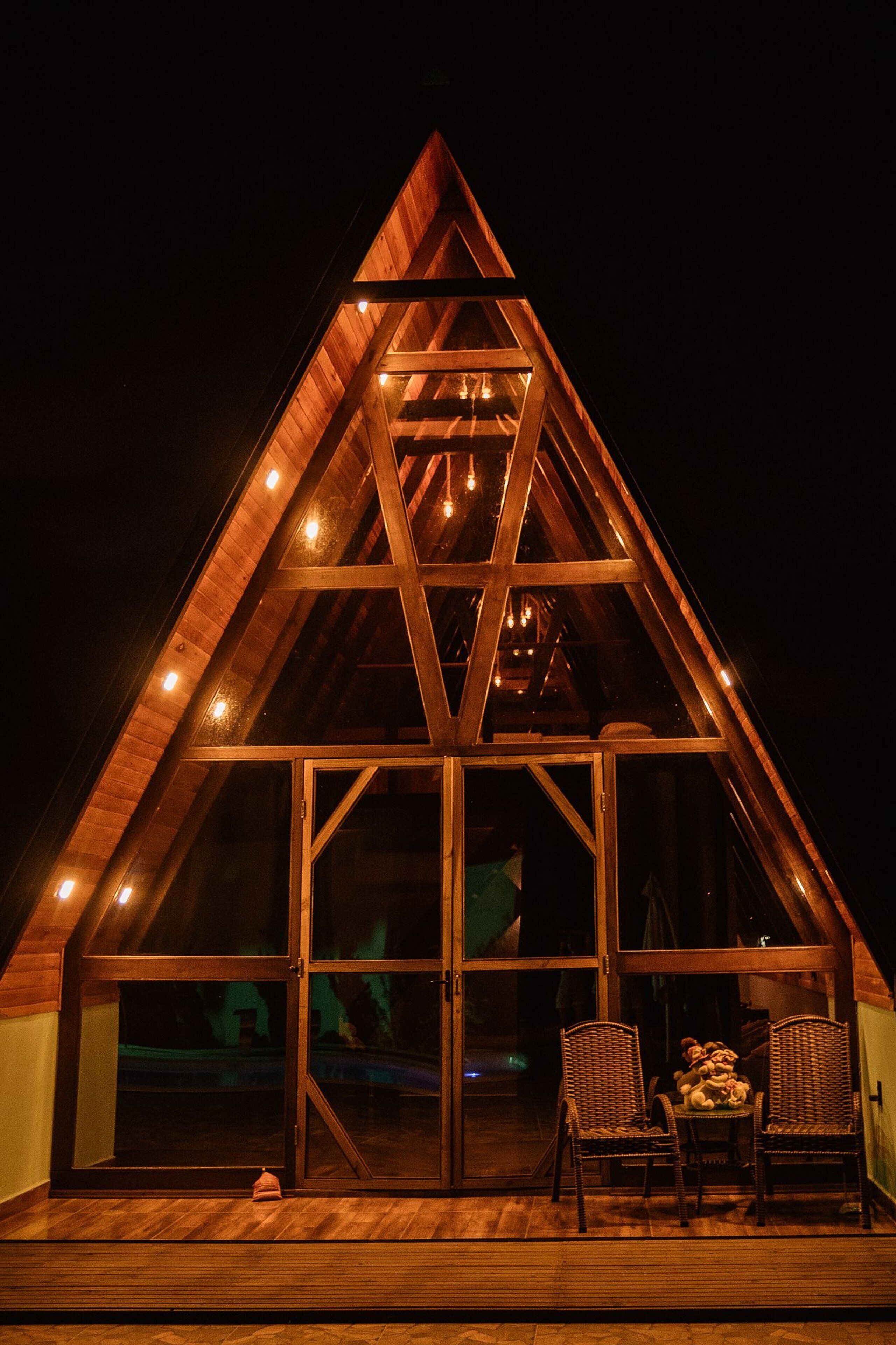 A triangular wooden structure with large glass windows and a wooden deck with two wicker chairs.