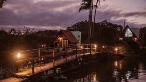 Eine Holzbrücke führt über ruhiges Wasser zu einem kleinen Haus mit rotem Dach.