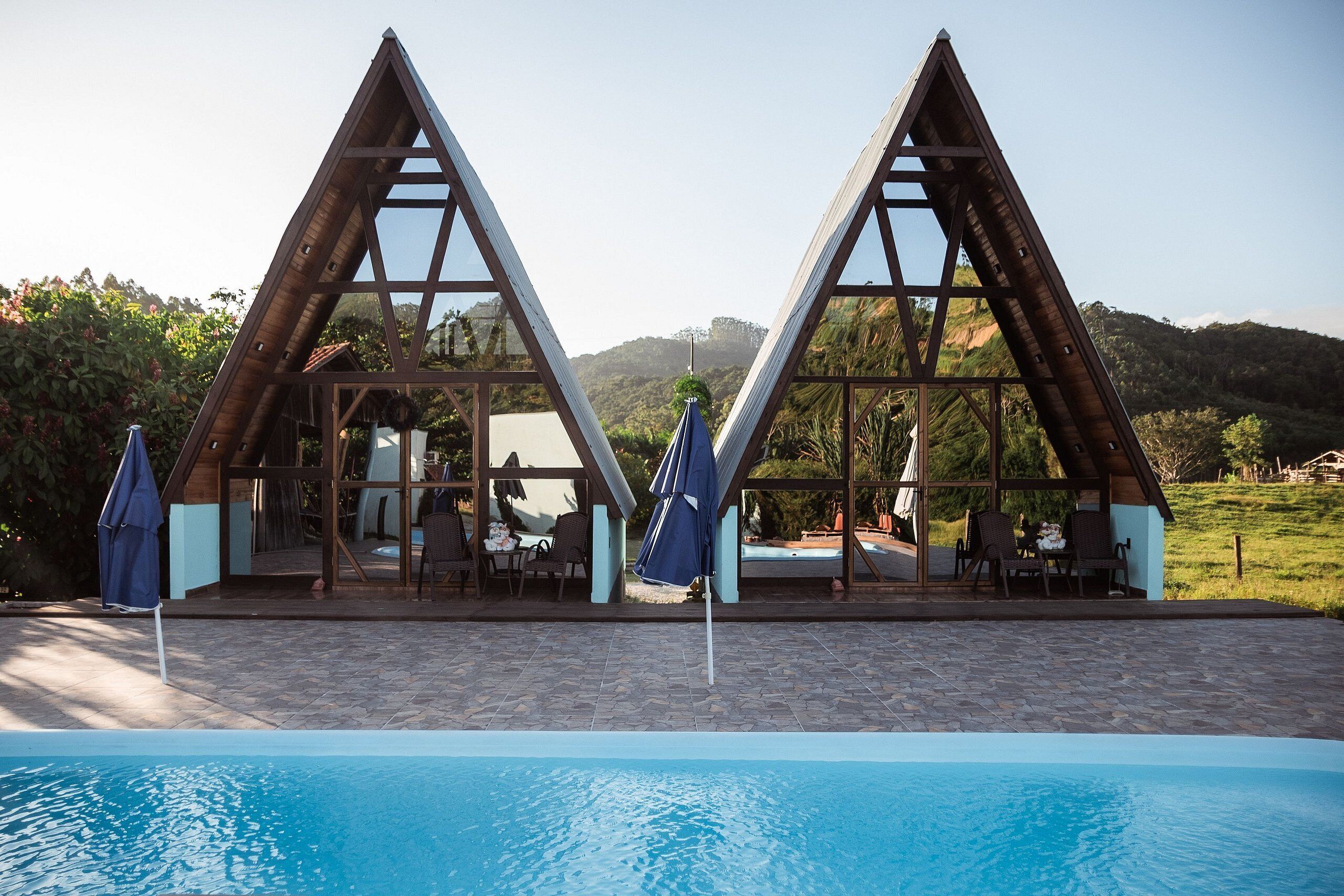 A poolside area with a wooden structure featuring triangular frames and glass windows, surrounded by lounge chairs and umbrellas.