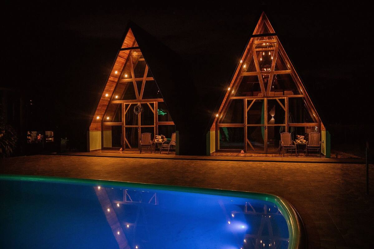 Two triangular wooden structures with glass windows, each with a pool table and chairs, adjacent to a swimming pool.