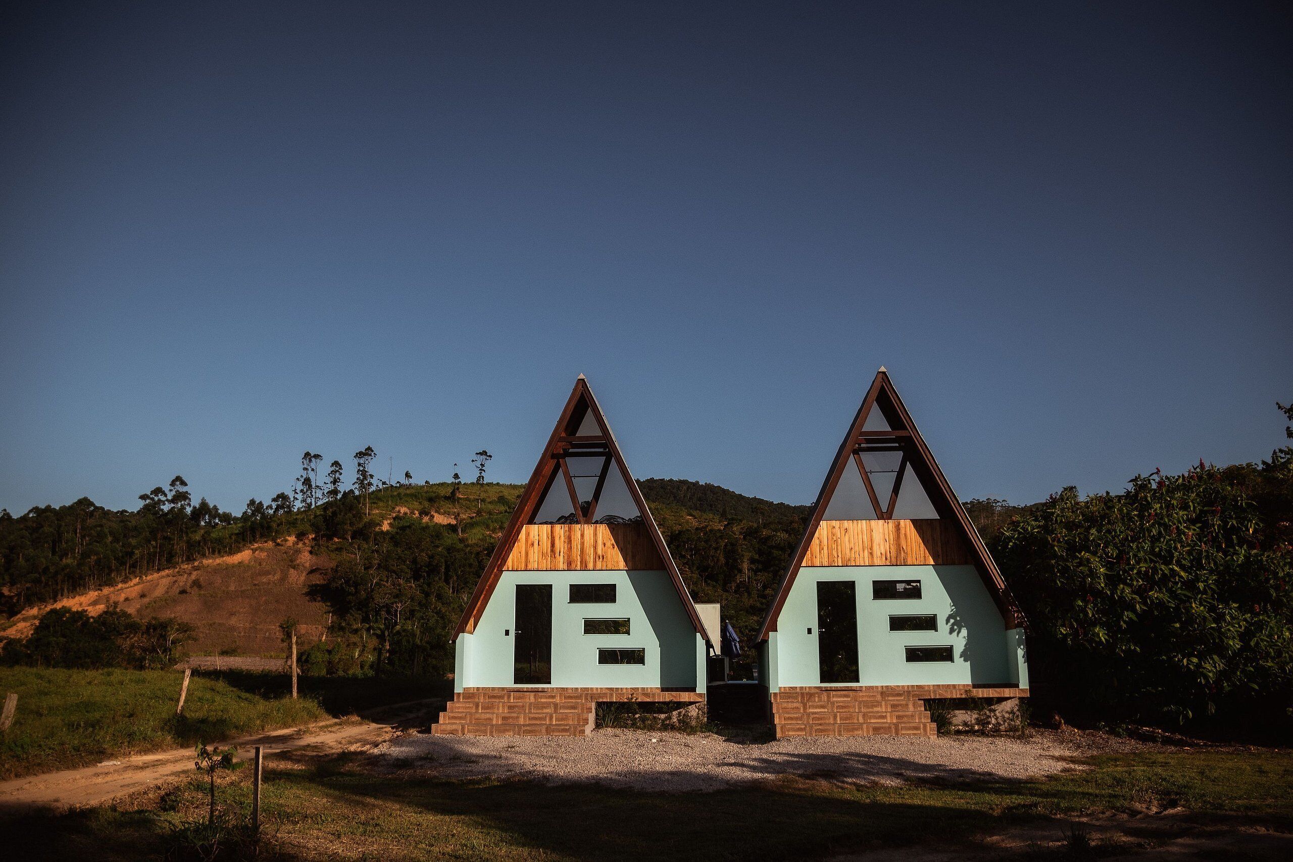 Dos cabañas de madera triangulares sobre base de ladrillo, ubicadas en un paisaje rural con cielo despejado.