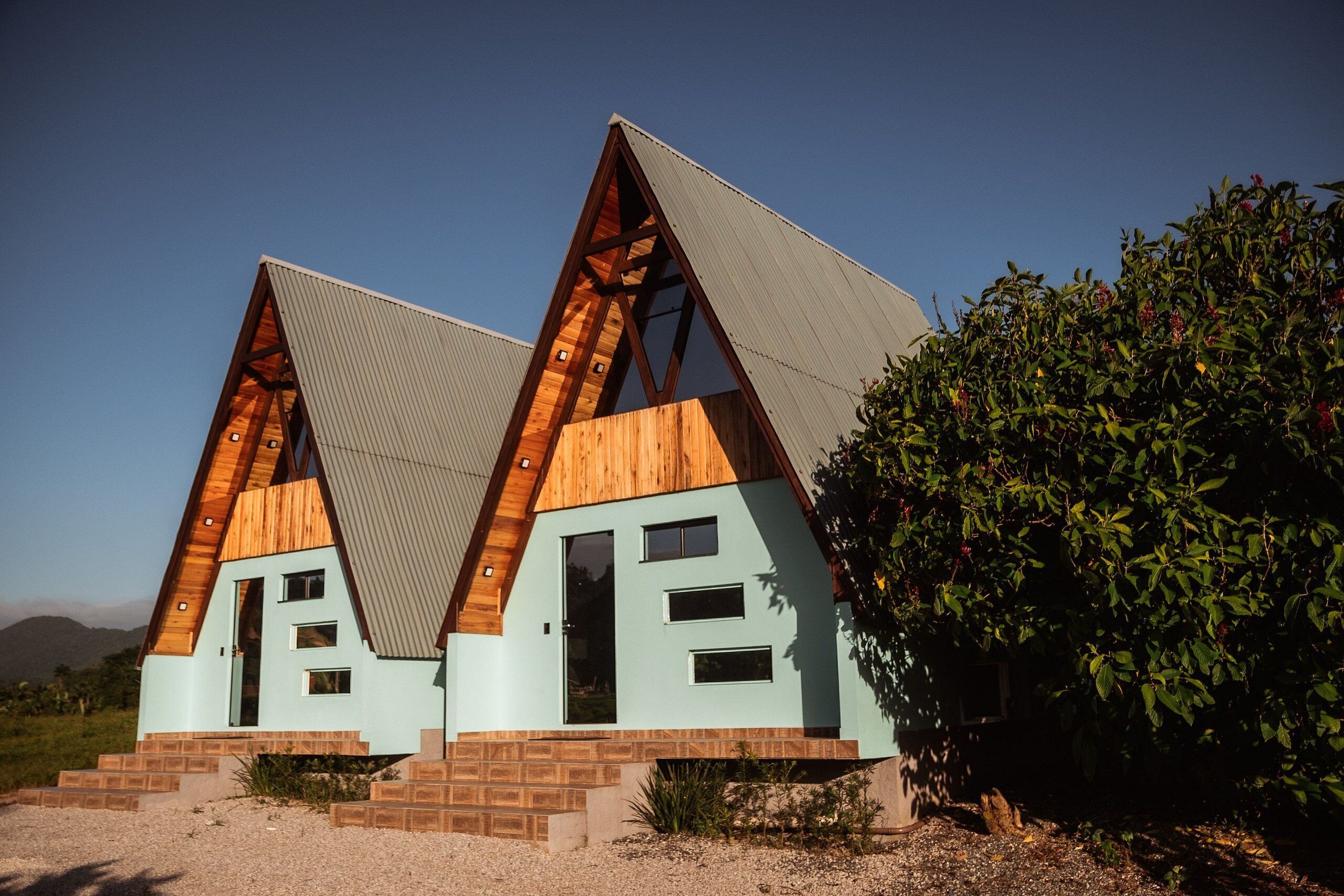 Un edificio moderno de forma triangular con fachada de madera y techo metálico, rodeado de grava y vegetación.