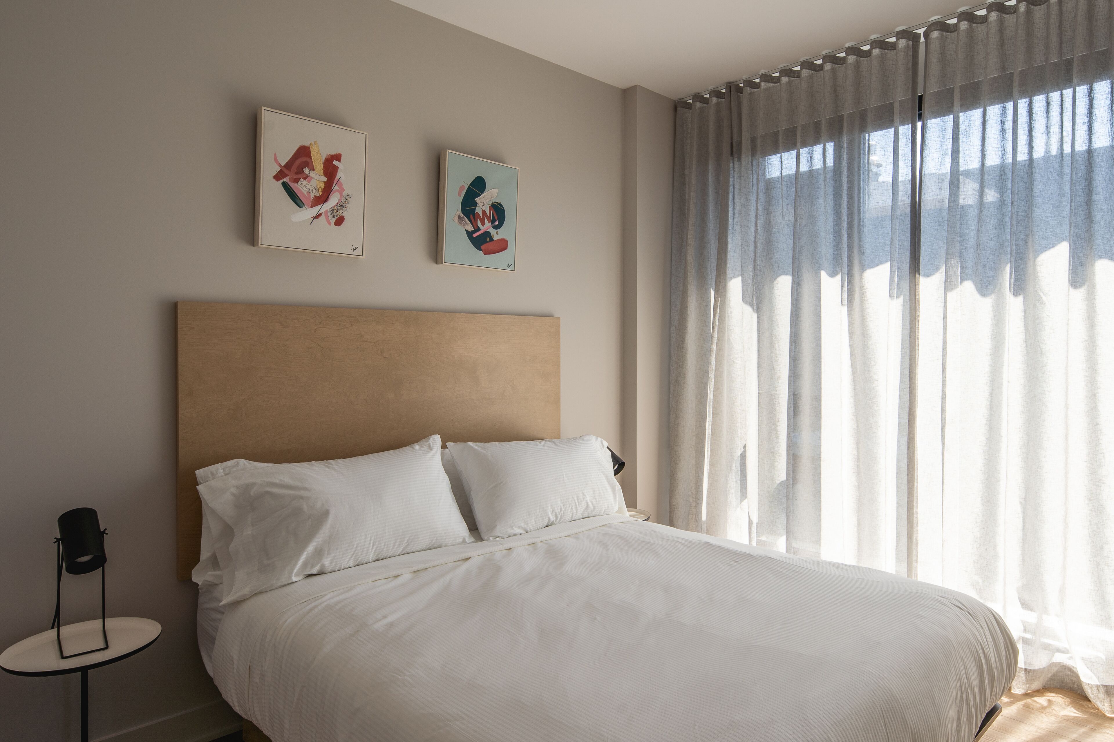 Una cama bien tendida con sábanas blancas, una mesita redonda con lámpara y dos cuadros enmarcados en la pared.