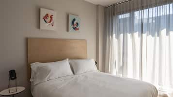 Una cama bien tendida con sábanas blancas, una mesita redonda con lámpara y dos cuadros enmarcados en la pared.