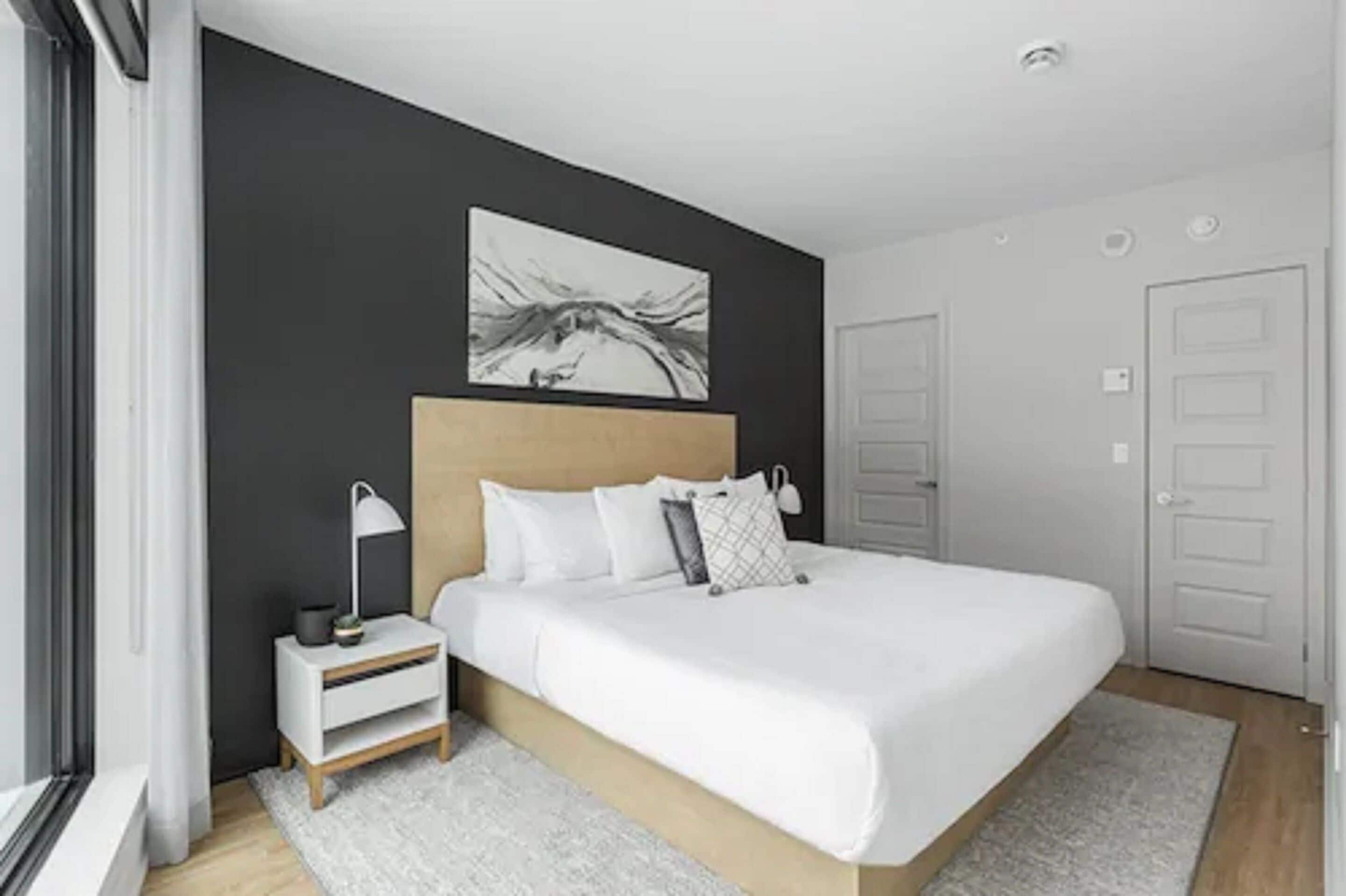 A modern bedroom with a large bed, a bedside table with a lamp, a painting on the wall, and a door to the right.