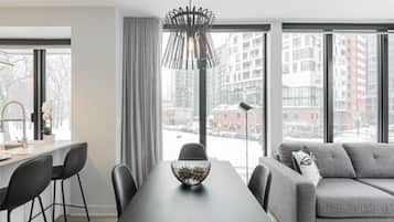 A modern living room with a dining table, grey sofa, and large windows offering a city view.
