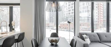 A modern living room with a dining table, grey sofa, and large windows offering a city view.