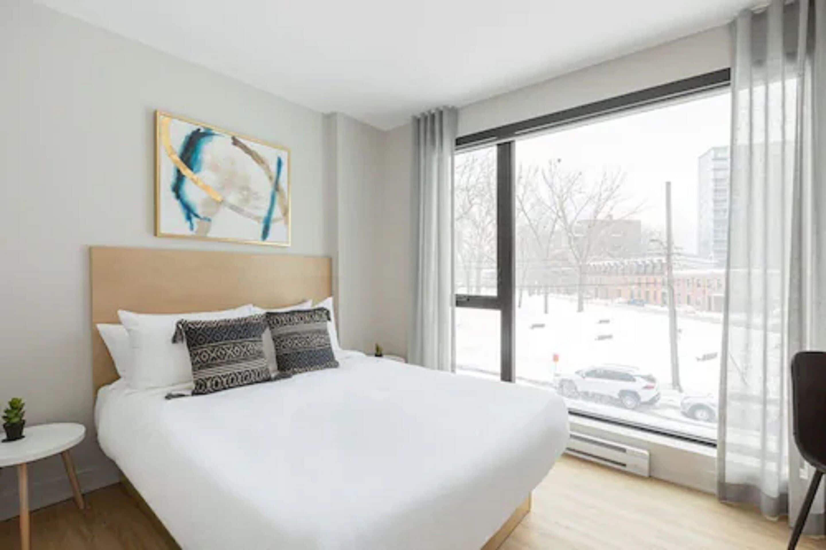 A modern hotel room with a large bed, a small table with a plant, a window with a view of the city, and a painting on the wall.