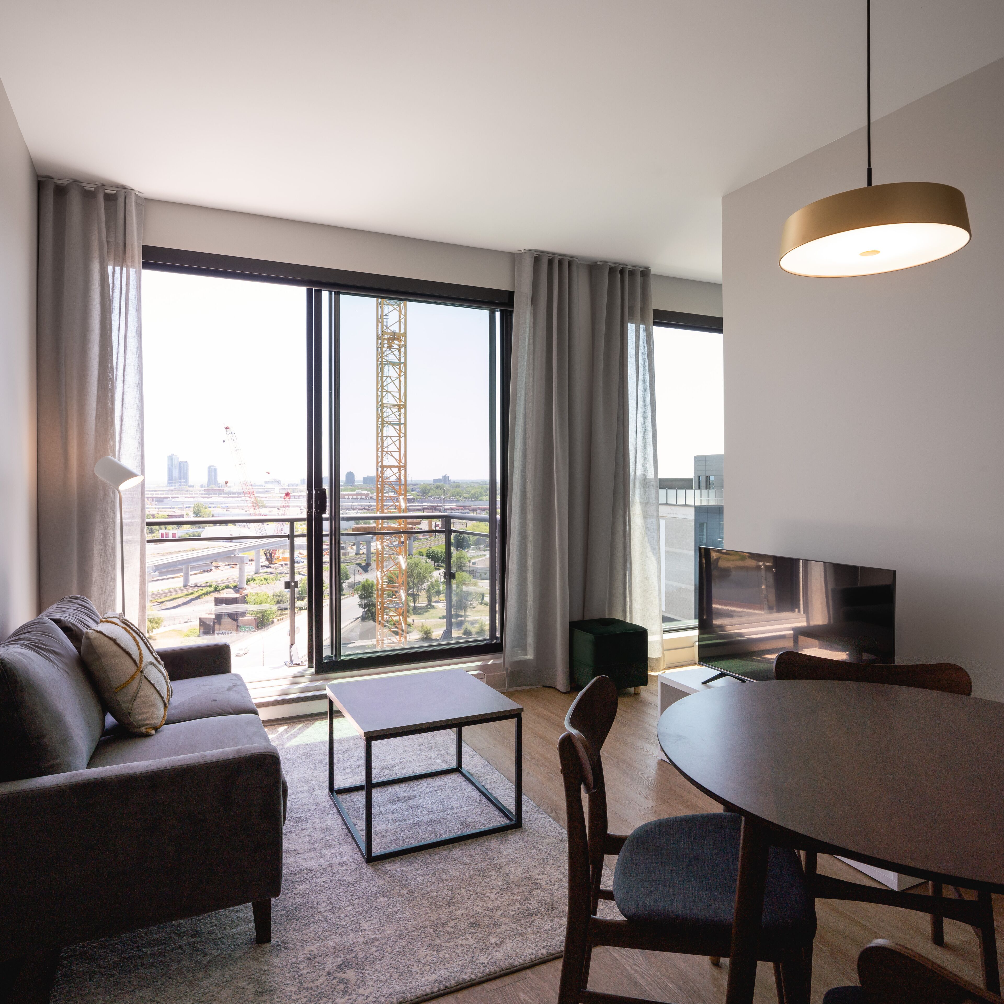Ein modernes Wohnzimmer mit einer Couch, einem Esstisch und einem Fernseher. Große Fenster bieten Blick auf eine Baustelle und die Stadtlandschaft.
