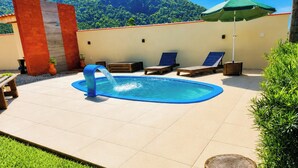 A swimming pool with a water feature, surrounded by lounge chairs and a green umbrella, with a mountain backdrop. - Sítio Morada do Pai com Sinuca e Piscina - Canelinha - SC (Canelinha)