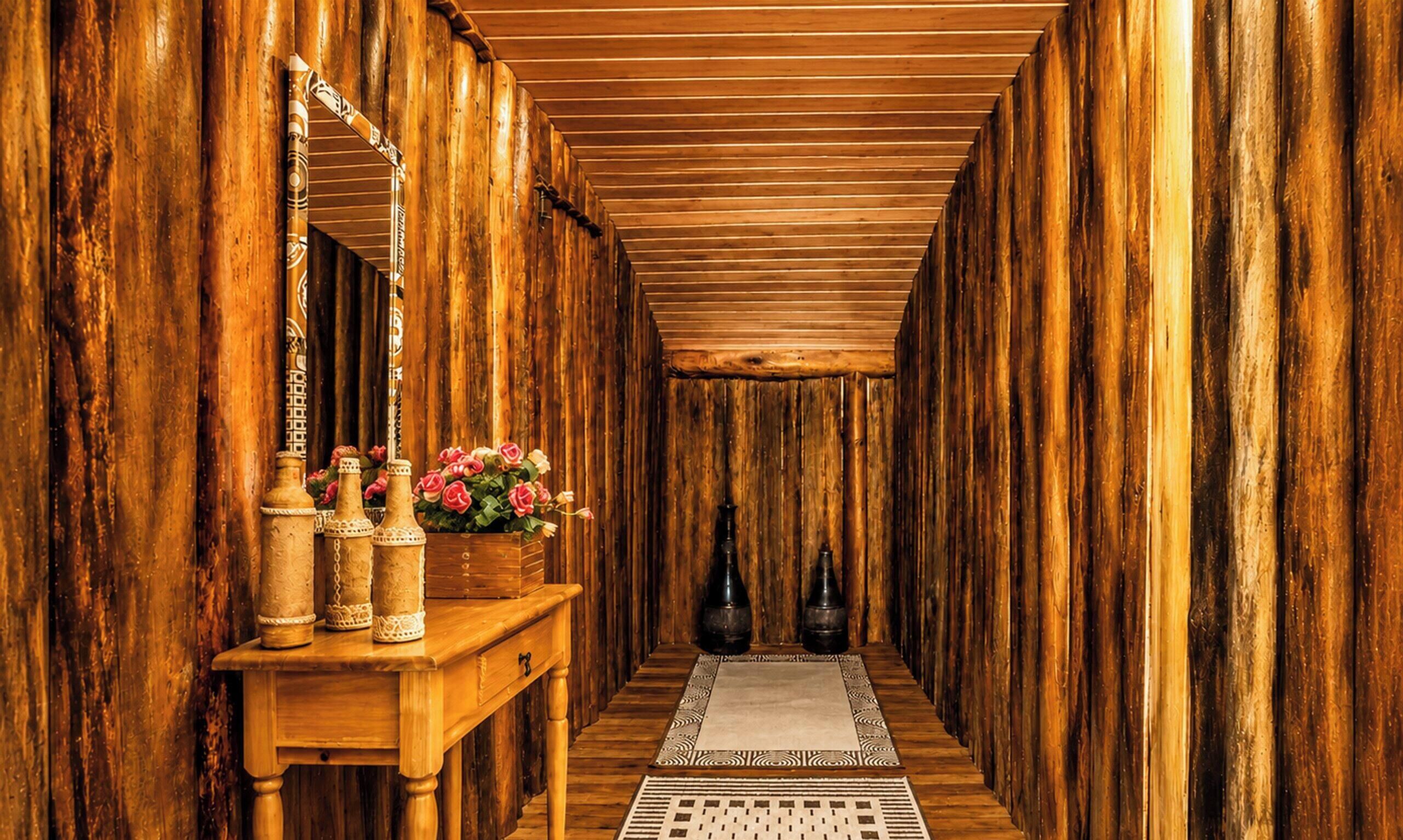 A narrow hallway with wooden walls and a wooden ceiling, leading to a room. There is a wooden bench with decorative vases and a potted plant with flowers.
