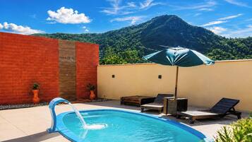 A swimming pool with a water feature, two lounge chairs, an umbrella, and a mountain view in the background.