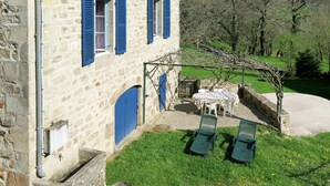 Un edificio de piedra con contraventanas azules, un patio con mesa y sillas, y un jardín con un pérgola.