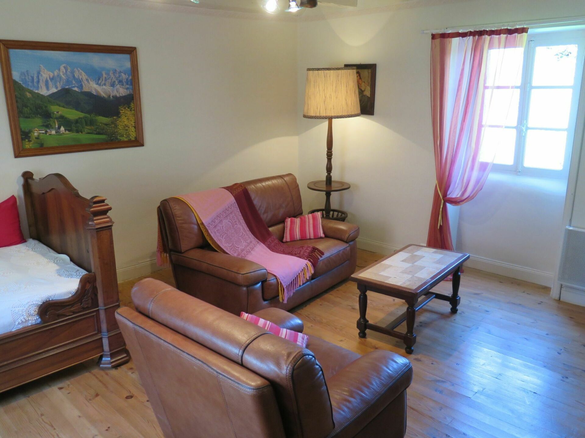 A living room with a brown leather sofa, a wooden table, a lamp, a bed with a wooden headboard, and a painting of mountains on the wall.