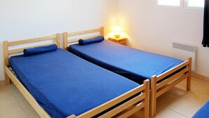 A small, simple room with two wooden beds, a nightstand with a lamp, and a window. - Cosy apartment for 6 guests with TV, terrace and pets allowed (Carcans-plage)