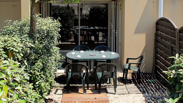 Un patio con mesa y sillas, rodeado de follaje y una terraza de madera.