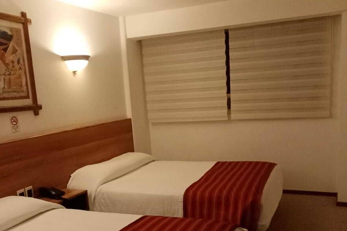 Une chambre d’hôtel avec deux lits, une table de chevet, une lampe fixée au mur et une fenêtre avec des stores.