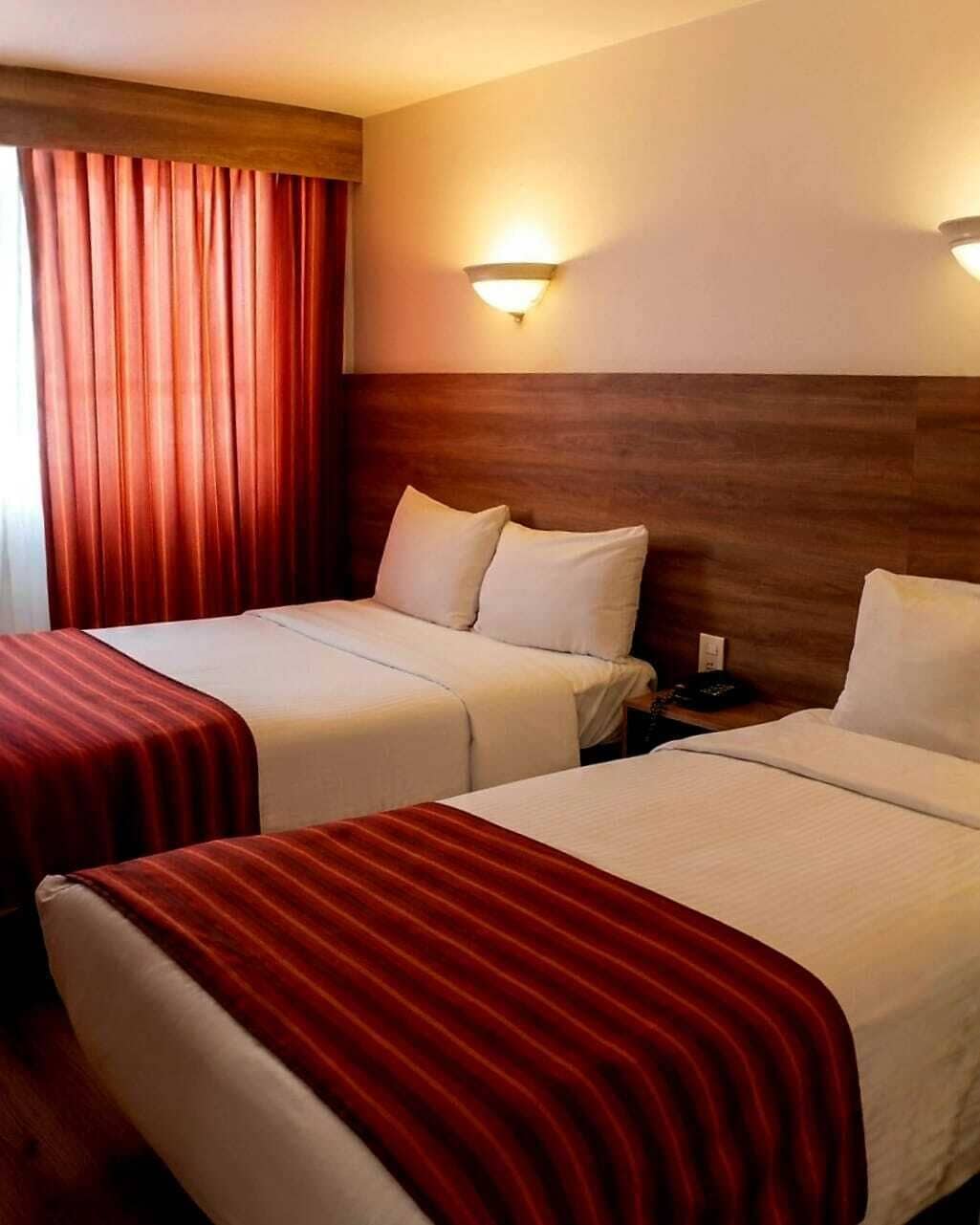 A hotel room with two beds, a wooden headboard, a red-striped bedspread, and a wall-mounted light fixture.