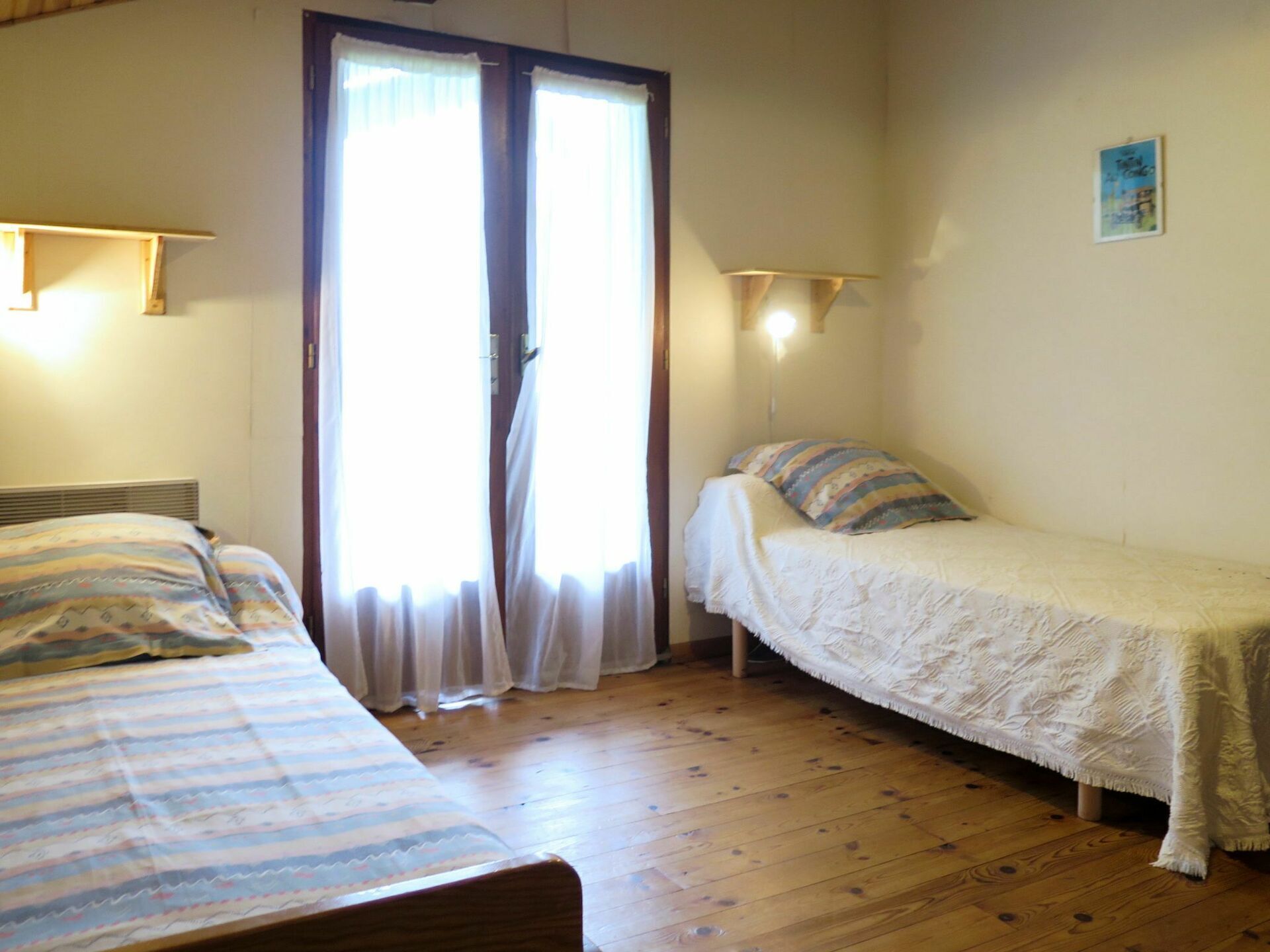Una habitación con dos camas, suelo de madera, una puerta corredera de vidrio y una lámpara pequeña fijada a la pared.