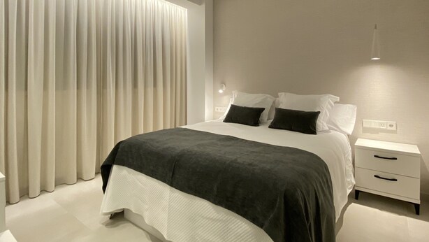 Un dormitorio moderno con una cama grande, cortinas blancas y una mesita de noche con lámpara.