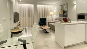 Una sala moderna con una mesa de comedor de vidrio, un sillón azul, un sofá, una mesa de centro y una zona de cocina con un microondas.