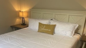A neatly made bed with white bedding and a 'Welcome' pillow, a bedside table with a lamp, and a wooden floor. -  NEAR UO, NEW RENO, SLPS 8, PET STAYS FREE, DECK/GRILL, FENCED YARD, VERY CLEAN
 (Eugene)