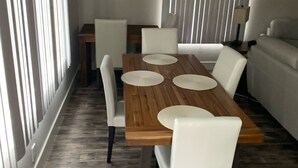 A dining area with a wooden table and white chairs, air conditioning unit on the wall, and a window with blinds. -  NEAR UO, NEW RENO, SLPS 8, PET STAYS FREE, DECK/GRILL, FENCED YARD, VERY CLEAN
 (Eugene)